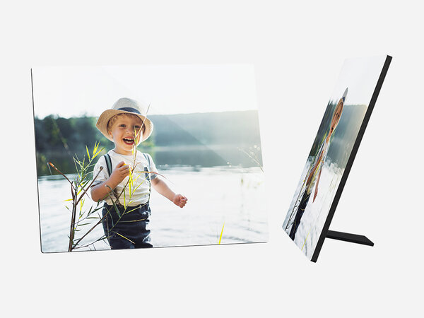 Ein quadratischer Fotoaufsteller zeigt ein fröhliches kleines Kind, das am Ufer eines Sees oder Flusses steht. Das Kind hat blonde Haare und trägt einen hellen Hut, ein weißes T-Shirt mit einem bunten Aufdruck und dunkle Latzhosen.